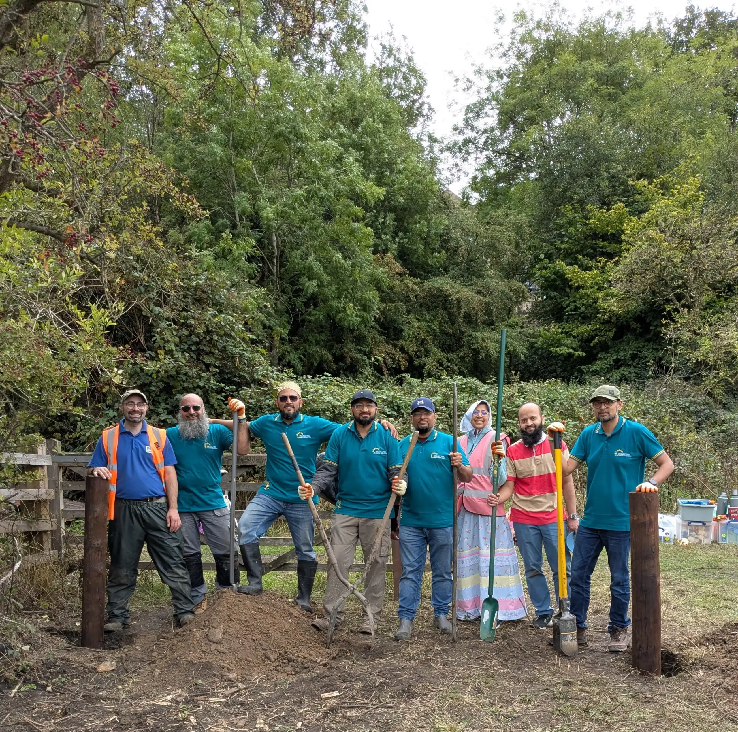 Seven men and one woman standing in a field holding gardening tools, surrounded by trees.