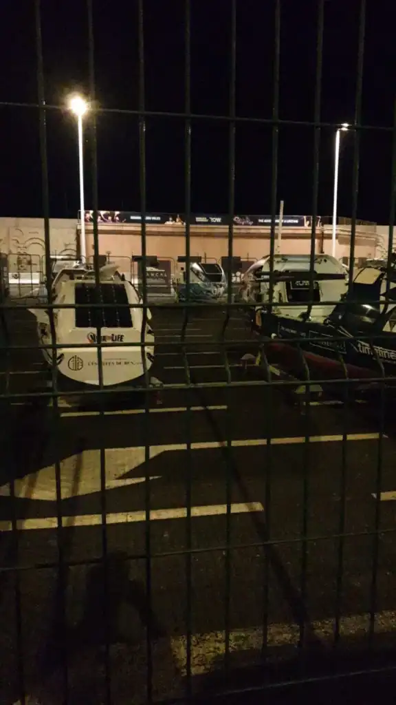 Urban river park at night with parked vehicles, fencing, and streetlights illuminating the area.